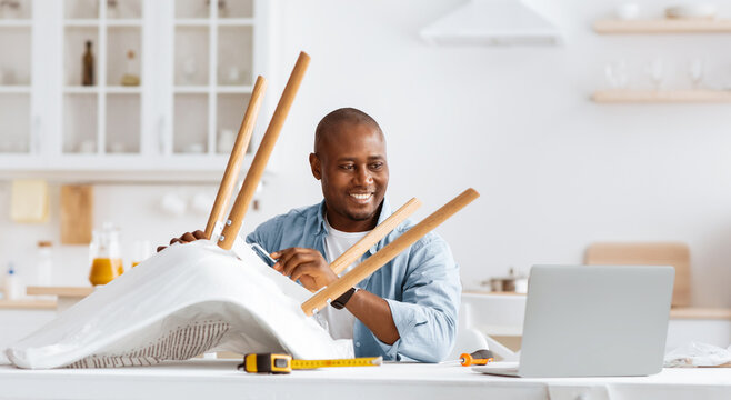 Happy african american man twisting part in chair and looking at laptop, watching online video instructions or manual in blog, sitting in kitchen interior, copy space. Homework and furniture assembly