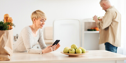 App for ordering food, delivery groceries at home during pandemic. Happy middle aged male with eco bags, take out vegetables in refrigerator, wife reads message in modern kitchen interior, copy space