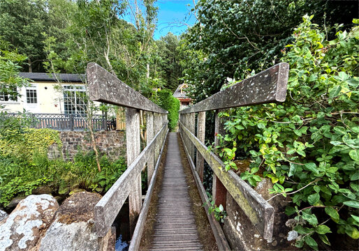 "Rustic bridge over Harden Beck leads to a cottage nestled in lush greenery. Blue sky and deep greens evoke a tranquil countryside retreat in Harden, Keighley."