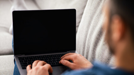 Over shoulder shot of man resting on couch in living room, using newest laptop with blank screen, websurfing or checking social media, chatting with friends, mockup, copy space © Prostock-studio