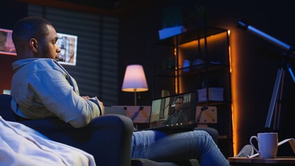 Focused black guy smiling and waving during a web conference in his living room, showcasing online...