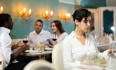 Single woman eating and drinking at the restaurant with a multiracial group of people on the background