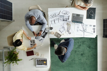 Top view of multiethnic group of young adult men and middle aged woman collaborating on architectural project, discussing blueprints and using laptop at shared workspace
