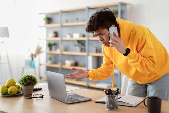 Confused Discontented Asian Man Talking On Phone Using Laptop Having Problem With Computer Or Internet Connection At Home. Displeased Male Customer Calling To Hotline Service And Complaining