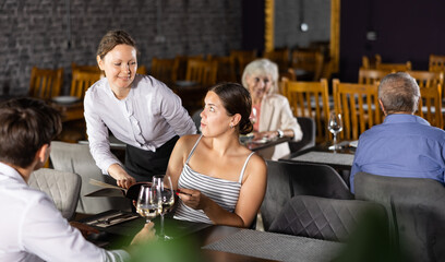 Polite smiling waitress talking to positive young couple visiting restaurant for romantic dinner, recommending dishes from menu card