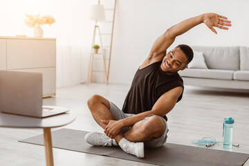 Online Workout. Cheerful Athletic African American Guy Doing Stretching Exercise At Laptop Training Sitting On Gymnastics Mat In Living Room At Home. Male Fitness Routine Concept