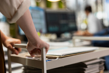 Close up of secretary in a modern office pushes a trolley with folders and files, providing...