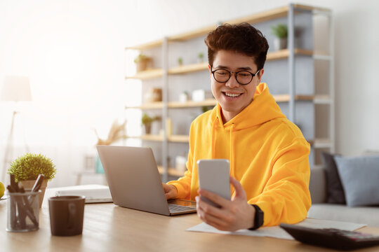 Business And Telecommuting Concept. Portrait of smiling young asian man using smartphone and modern laptop computer, reading text message, sitting at desk with gadgets, working at home office - Powered by Adobe