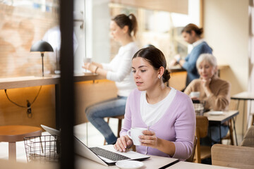 Smiling young woman using laptop and drinking coffee in modern cafe