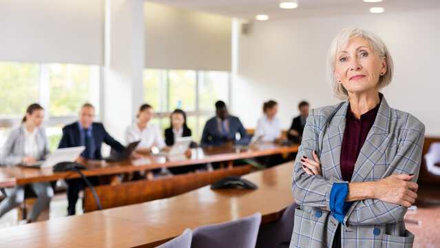 Smiling classy elegant senior mature white businesswoman on foreground posing at camera with arms crossed and office employees at back