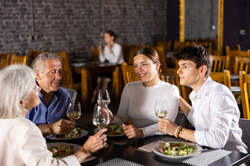 Happy young girl and guy meeting with elderly parents over dinner in restaurant. People talking cheerfully and drinking wine at table