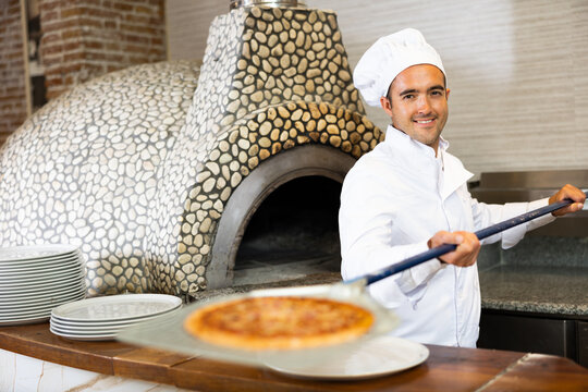 Portrait of smiling pizza chef at work, taking pizza from oven at restaurant kitchen