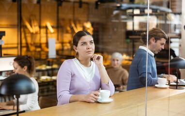 Dreamy attractive young female office worker sitting alone at table in cozy cafe, drinking fragrant...
