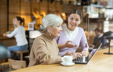 Friendly smiling young girl helping interested elderly lady to browse emails or websites on internet on laptop over cup of coffee in cozy cafe
