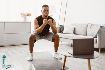 Domestic Workout. Athletic African American Guy Doing Deep Squats At Laptop During Online Training...