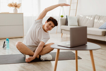 Positive athletic asian man stretching arms at home, using laptop, watching sport videos on...
