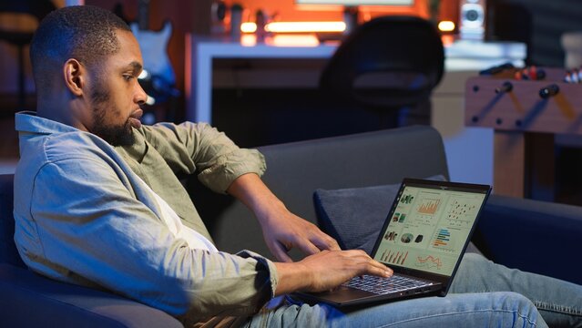 Black male trader monitoring stock market real time changes on laptop, investing funds with the goal to boost earnings. Focused young guy studying economic values for global advancement. Camera B.
