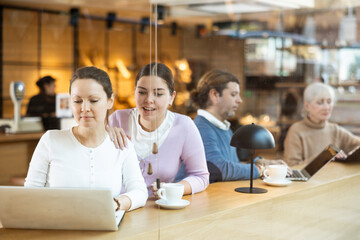 Portrait of two positive young females surfing Internet on notebook while drinking tea or coffee in cafeteria