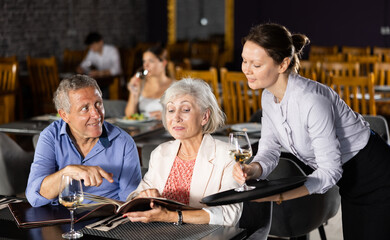 Adult female waiter in uniform takes order from elderly couple in restaurant