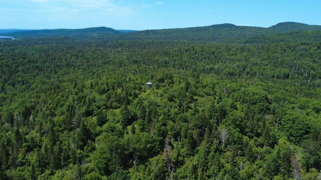 Panoramic aerial shot of an observation tower perched atop a lush, green mountain forest. Seigneurie du Lac Matapedia, Quebec, Canada, 2025.