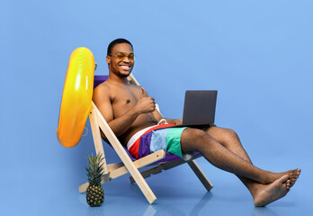 Cheerful black guy in swimwear using laptop, browsing internet during beach vacation, sitting in...