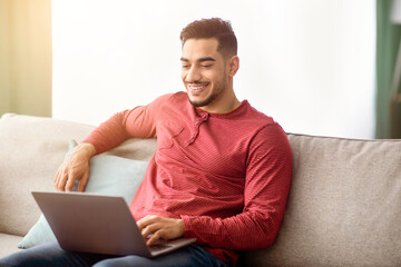 Cheerful arab guy sitting on couch at home, using laptop and smiling, watching movie online, copy...