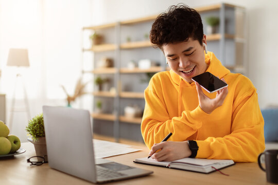 Work From Home. Young Asian Guy Talking On Mobile Phone And Taking Notes, Using Voice Assistant, Writing In Notebook. Smiling Man Sitting At Desk With Laptop At Home Office, Wearing Earbuds