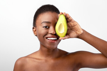 Happy african american young woman holding fresh avocado halves and covering eye, smiling to camera...