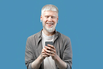 People and technology concept. Excited albino man using smartphone, chatting online or browsing social media, standing isolated over blue studio background and smiling to camera