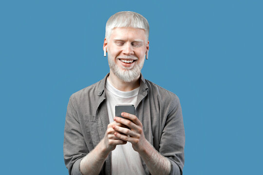 People and technology concept. Excited albino man using smartphone, chatting online or browsing social media, standing isolated over blue studio background and smiling to camera - Powered by Adobe
