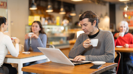 In cozy cafe hall, freelance man works with laptop and drinks coffee. Remote work from anywhere in world