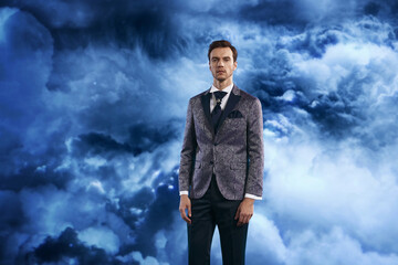Confident man in a stylish patterned suit jacket and dark trousers posing against a dramatic cloudy blue sky, showcasing modern elegance, menswear fashion, and lifestyle portrait photography.