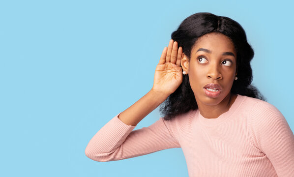 Curious young black female with hand near ear trying to overhear something, interested millennial african woman eavesdropping for fresh gossips, standing isolated over blue background, copy space