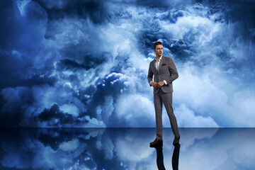 Obraz premium Confident man wearing a tailored light gray suit with a red pocket square, standing against a dramatic cloudy blue sky backdrop, symbolizing men’s fashion, modern elegance, lifestyle and photography.