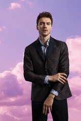 Confident man in a dark patterned blazer and shirt standing against dreamy pink clouds, perfect for modern men’s fashion, editorial style, and artistic portrait photography.