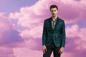 Stylish man wearing a green patterned blazer and dark pants standing against dreamy pink sky clouds, perfect for modern fashion and editorial portraits.