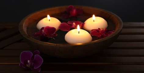 Burning candles and orchid flowers in bowl of water on wooden table against black background, closeup