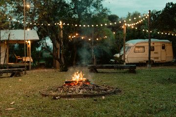 Cozy night camping in nature with glowing bonfire, fairy lights, parked caravan creating a peaceful...