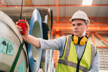 Professional engineer working in metal factory inspecting steel coil rolls, wearing safety gear,...