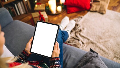 Woman holding tablet on couch in cozy living room decorated for Christmas, enjoying holiday season with warm and festive atmosphere.