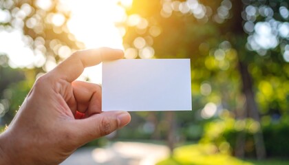 Hand holding blank white business card outdoors in sunny park setting, presenting opportunity and connection with a hopeful, professional tone.