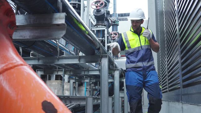 Industrial engineering technician worker working maintenance building exterior plumbing systems. Male engineer in safety uniform speaking on radio and checking pipeline valves at construction site.