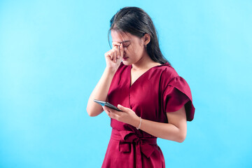 A young woman in a red dress holds a smartphone while closing her eyes and touching her face,...