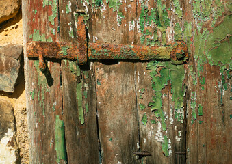 A rustic latch is covered in rust, set against a wooden door with peeling green paint. The texture of the wood and the aged metal creates a visually compelling contrast. 
