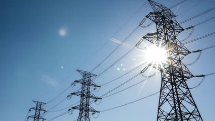 Towering electricity pylons stretch across a clear blue sky, with the bright sun shining through the power lines - Powered by Adobe