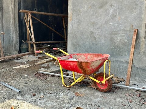 Red wheelbarrow (gerobak sorong) with an unfinished house in the background. August 8, 2025 - Depok, Indonesia.