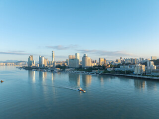 Naklejka premium Coastal Xiamen China cityscape reflects morning light on tranquil water as boats glide past modern waterfront buildings.