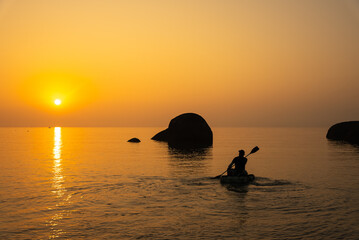 Golden sunset casts a warm glow on the calm ocean water as a kayaker paddles past large rock...