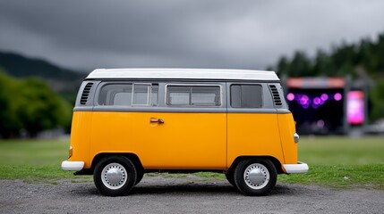 Yellow and grey vintage camper van parked on gravel near a blurred outdoor music festival stage