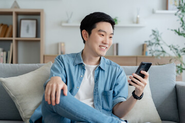 Smiling handsome young Asian man sitting on cozy couch, using funny mobile apps in living room. man...